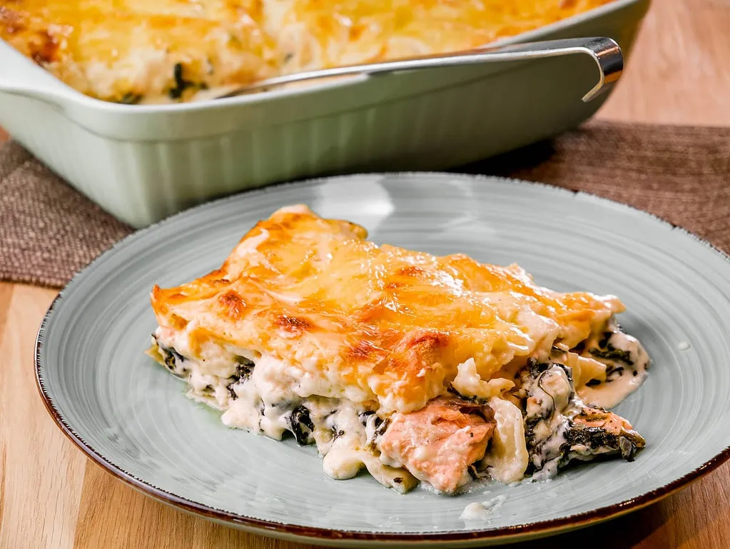 Ein Stück Lachs-Spinat-Lasagne mit einer goldbraunen Käseschicht, serviert auf einem Teller. Im Hintergrund befindet sich eine Auflaufform mit der restlichen Lasagne.