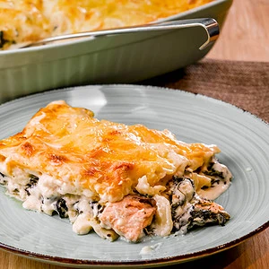 Ein Stück Lachs-Spinat-Lasagne mit einer goldbraunen Käseschicht, serviert auf einem Teller. Im Hintergrund befindet sich eine Auflaufform mit der restlichen Lasagne.