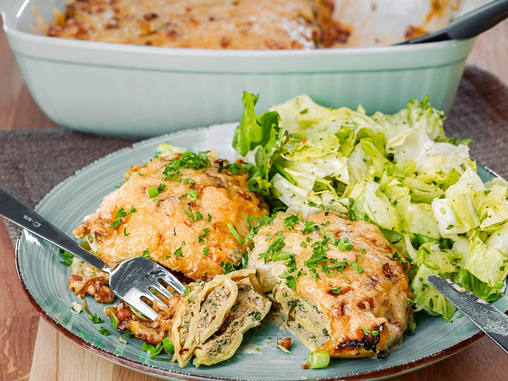 Ein Teller mit einer Portion überbackener Maultaschen, daneben ein frischer grüner Salat. Im Hintergrund ist eine Auflaufform mit dem restlichen Gericht zu sehen.