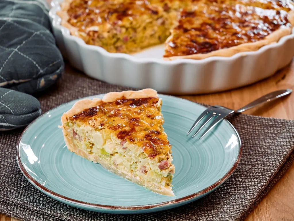 Ein Stück Quiche mit Lauch und Schinken, serviert auf einem Teller, der restliche Kuchen im Hintergrund.