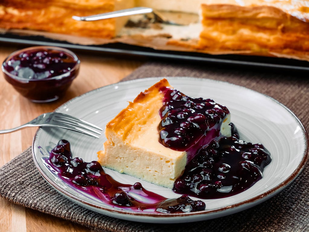 Ein Stück Käsekuchen mit Heidelbeersoße auf einem Teller, Heidelbeeren und Käsekuchen im Hintergrund.