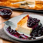 Ein Stück Käsekuchen mit Heidelbeersoße auf einem Teller, Heidelbeeren und Käsekuchen im Hintergrund.