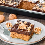 Ein Stück Zucchini-Nusskuchen mit Schokoladenglasur und Walnüssen, serviert auf einem Teller, restlicher Kuchen im Hintergrund.