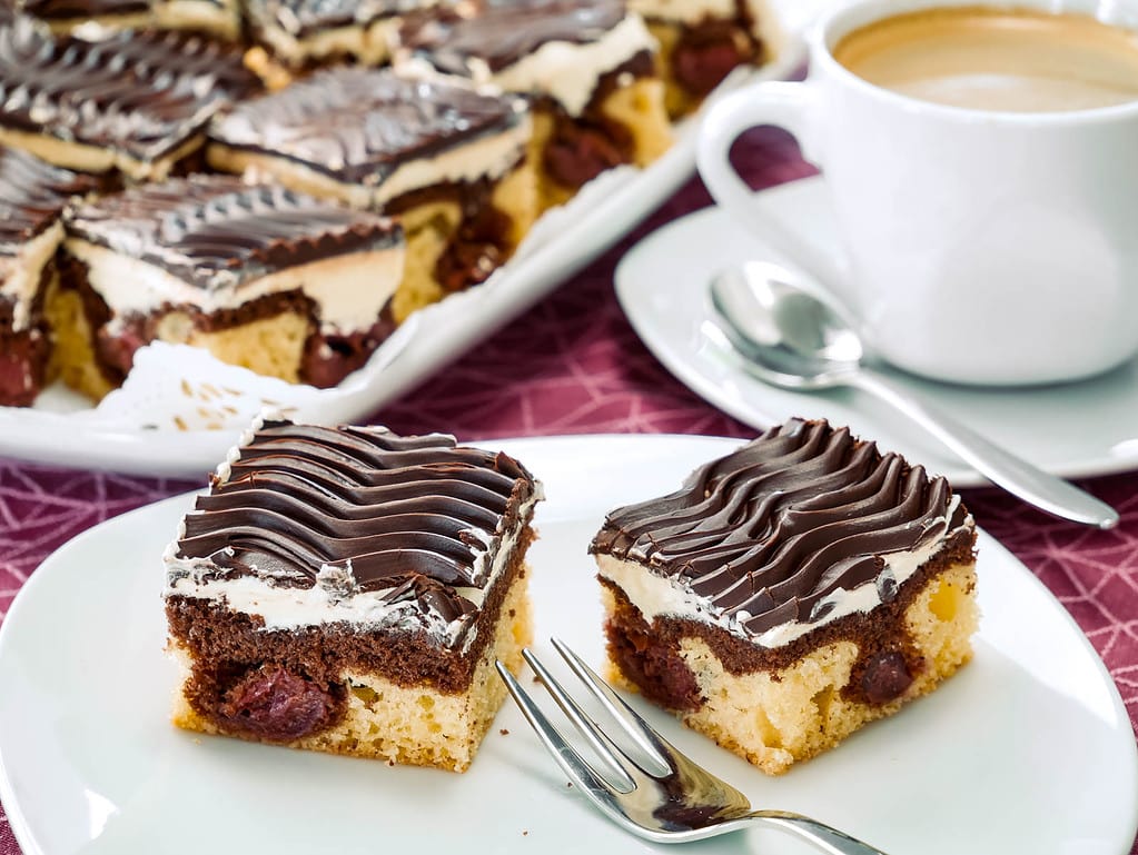 Mehrere Stücke Donauwelle, ein Marmorkuchen mit einer Schicht Kirschen, Buttercreme und Schokoladenglasur, serviert auf einem Teller. Im Hintergrund steht ein Tablett mit weiteren Stücken.