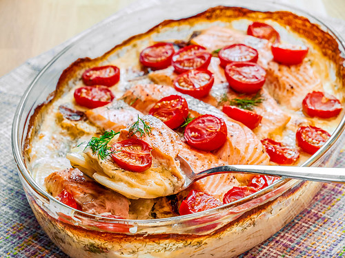 Ein Kartoffelgratin mit Lachsfilets, Kirschtomaten und einer cremigen Dill-Sahne-Soße, serviert in einer Glasform.