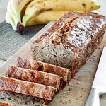 Ein frisch gebackenes Bananenbrot, in Scheiben geschnitten und auf einem Holzbrett serviert. Im Hintergrund liegen reife Bananen.