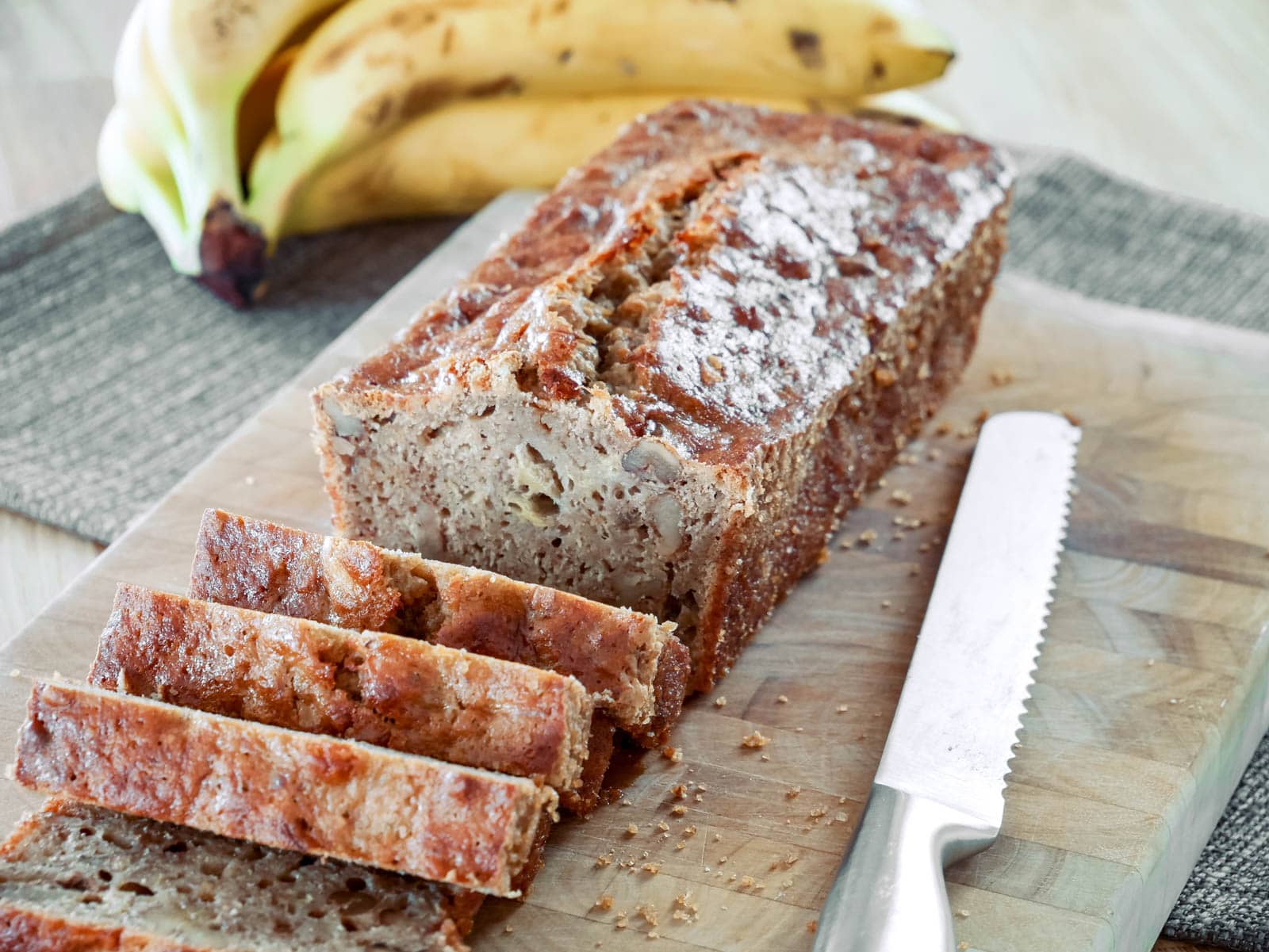 Ein frisch gebackenes Bananenbrot, in Scheiben geschnitten und auf einem Holzbrett serviert. Im Hintergrund liegen reife Bananen.