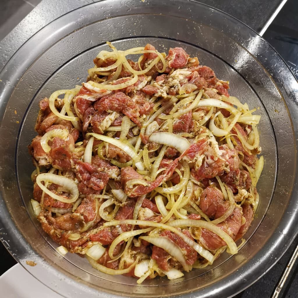 Mariniertes Gyrosfleisch, gemischt mit Zwiebeln, in einer Metallschüssel als Vorbereitung für den Gyros-Schichtbraten.