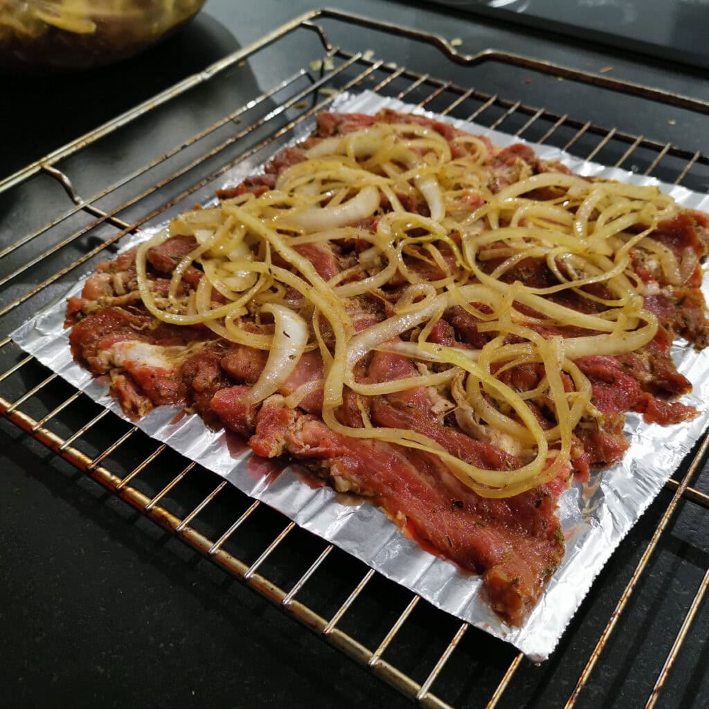 Eine einzelne Schicht rohes, mariniertes Gyrosfleisch auf einem Rost, belegt mit einer Schicht Zwiebeln.