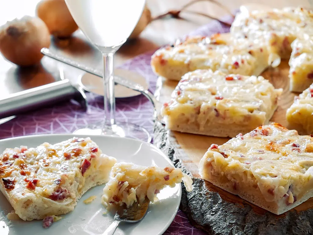 Ein Stück Zwiebelkuchen, belegt mit geschmolzenem Käse und Speck, serviert auf einem Teller mit einer Gabel.