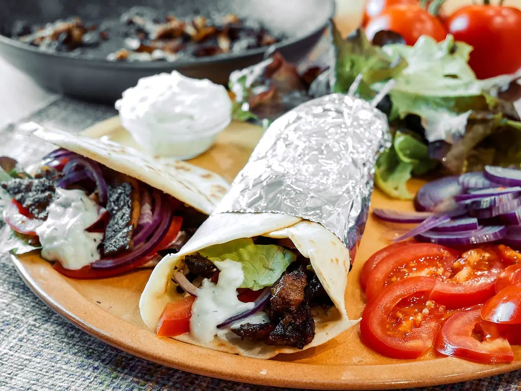 Ein Gyros-Wrap, teilweise in Folie eingewickelt, gefüllt mit Salat, Tomaten, Zwiebeln und Tsatsiki, serviert auf einem Teller mit frischem Salat, Tomatenscheiben und roten Zwiebeln. Im Hintergrund befindet sich eine Pfanne mit Gyrosfleisch und eine Schüssel mit Tsatsiki.