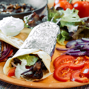 Ein Gyros-Wrap, teilweise in Folie eingewickelt, gefüllt mit Salat, Tomaten, Zwiebeln und Tsatsiki, serviert auf einem Teller mit frischem Salat, Tomatenscheiben und roten Zwiebeln. Im Hintergrund befindet sich eine Pfanne mit Gyrosfleisch und eine Schüssel mit Tsatsiki.