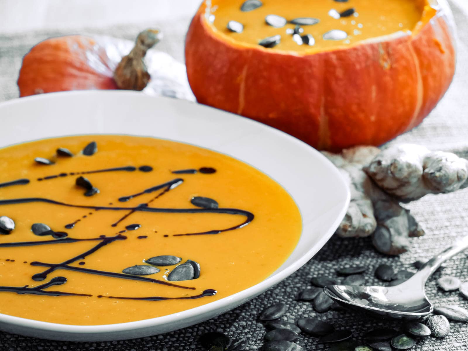 Ein Teller mit Kürbissuppe, garniert mit Kürbiskernen und einem Spritzer Kürbiskernöl. Im Hintergrund steht ein ausgehöhlter Kürbis, ebenfalls gefüllt mit Suppe und Kürbiskernen.