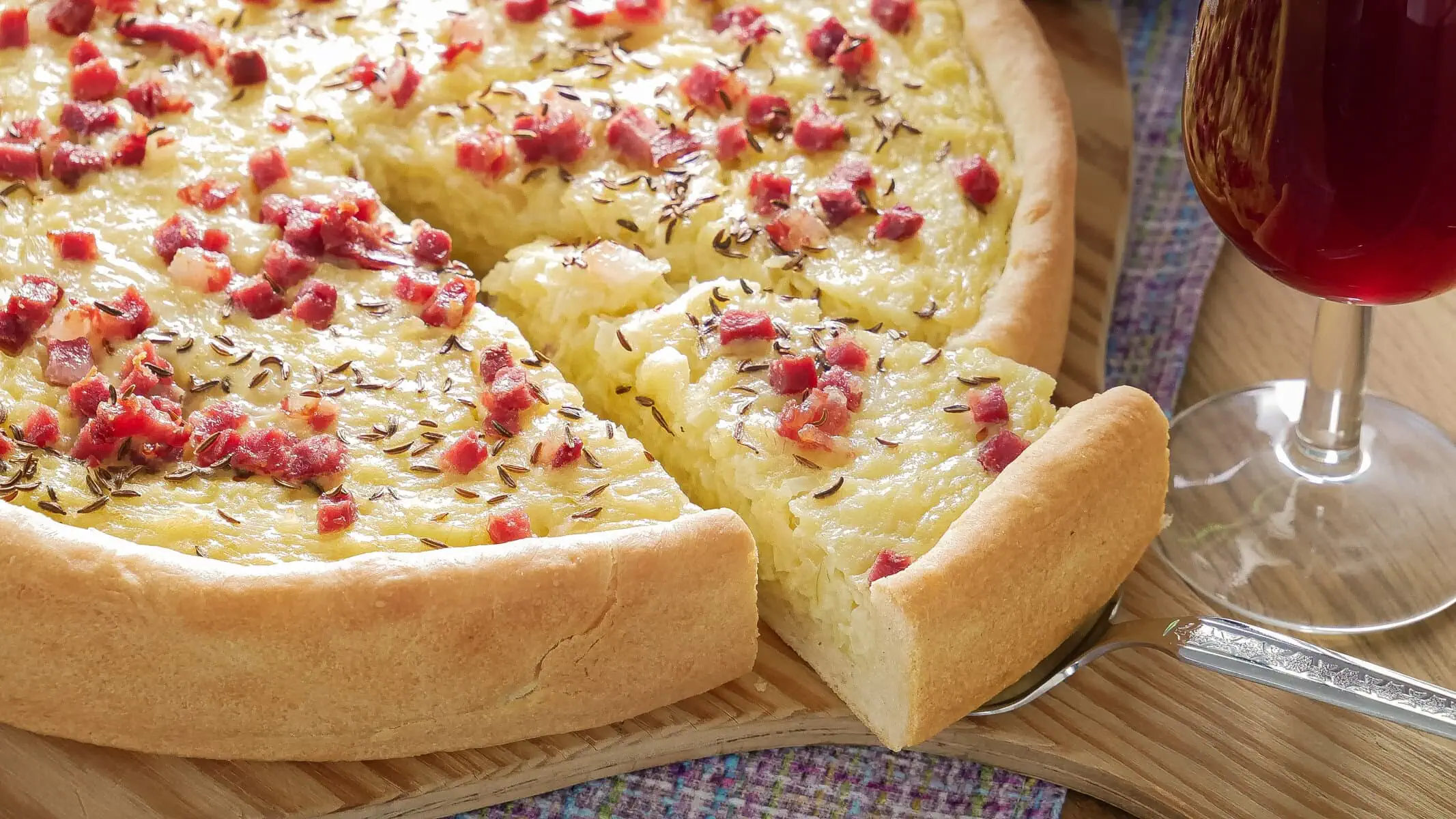 Nahaufnahme eines schwäbischen Zwiebelkuchens mit Speckwürfeln und Kümmel, ein Stück wurde herausgeschnitten.