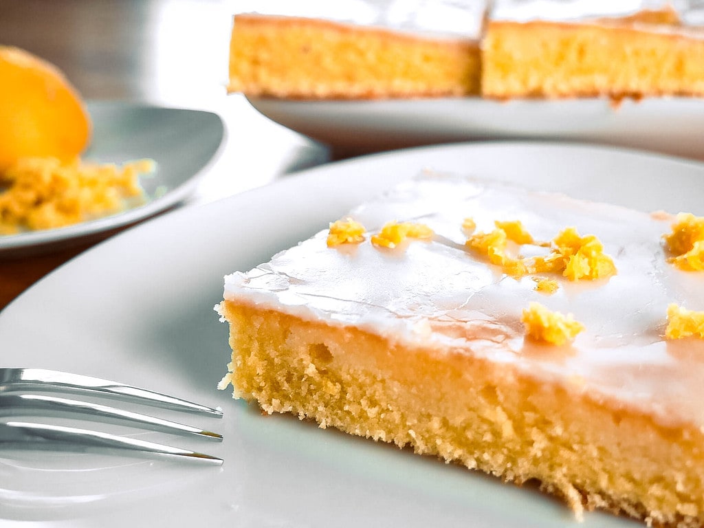 Ein Stück Zitronenkuchen mit Zuckerguss auf einem Teller, der restliche Kuchen im Hintergrund.