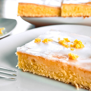 Ein Stück Zitronenkuchen mit Zuckerguss auf einem Teller, der restliche Kuchen im Hintergrund.