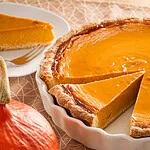 Ein angeschnittener Pumpkin Pie in einer weißen Tarteform, mit einem Stück auf einem Teller und einem Hokkaido-Kürbis im Vordergrund.