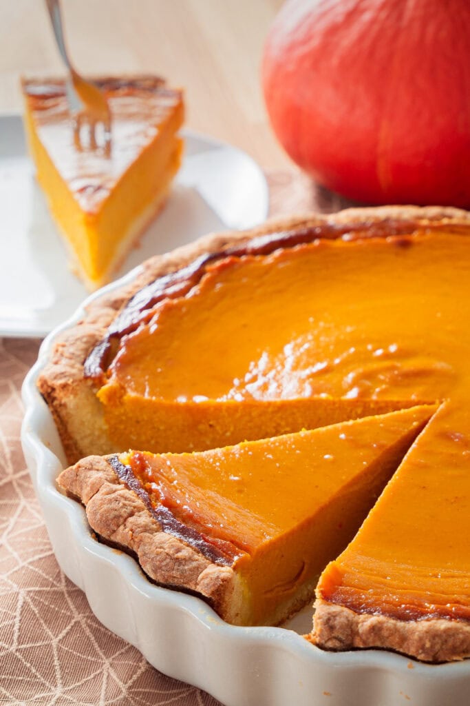 Ein Stück cremiger Pumpkin Pie auf einem Teller, mit einem Hokkaido-Kürbis und der restlichen Tarte im Vordergrund.