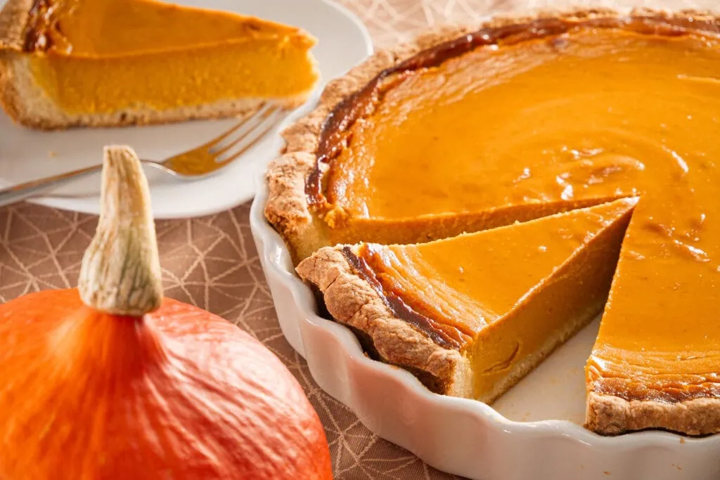 Ein Stück Pumpkin Pie auf einem Teller, mit einem Hokkaido-Kürbis im Vordergrund und der restlichen Tarte im Hintergrund.