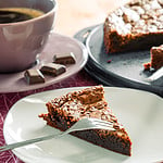 Ein Stück Tarte au Chocolat auf einem Teller, daneben eine Tasse Kaffee und einige Schokoladenstücke. Im Hintergrund steht der restliche Kuchen auf einer Kuchenplatte.