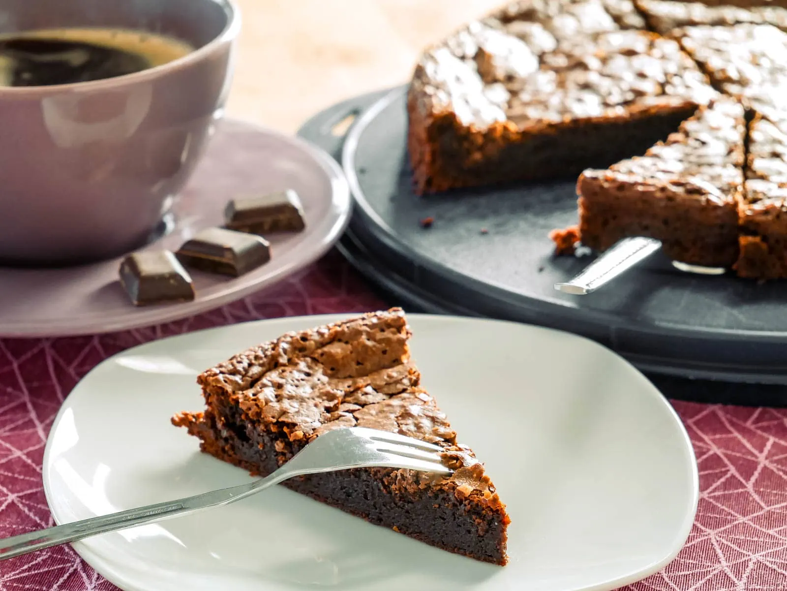 Ein Stück Tarte au Chocolat auf einem Teller, daneben eine Tasse Kaffee und einige Schokoladenstücke. Im Hintergrund steht der restliche Kuchen auf einer Kuchenplatte.