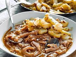 Schwäbische Saure Kutteln, serviert mit Bratkartoffeln auf einem Teller. Im Hintergrund ist eine Pfanne mit weiteren Bratkartoffeln zu sehen.