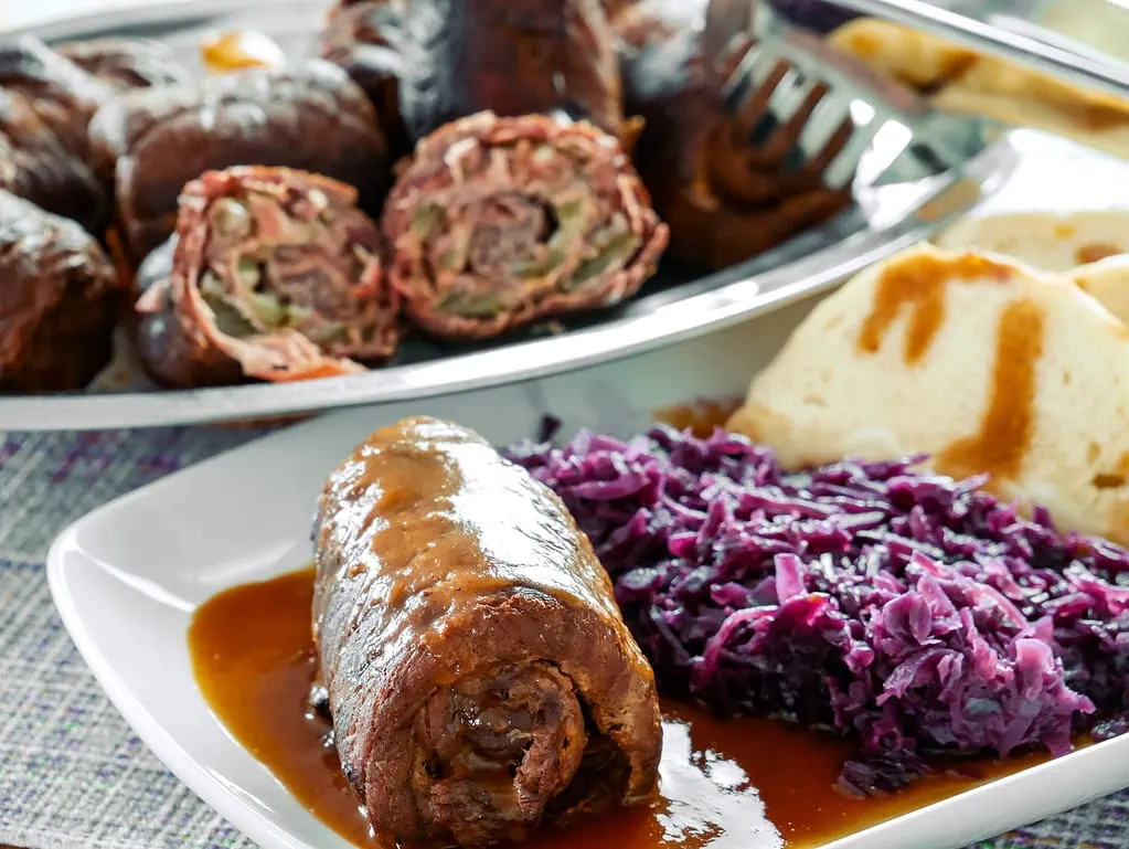 Rinderrouladen gefüllt mit Speck und Gurken, serviert mit Rotkohl, Knödeln und Bratensauce auf einem Teller.