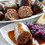 Rinderrouladen gefüllt mit Speck und Gurken, serviert mit Rotkohl, Knödeln und Bratensauce auf einem Teller.