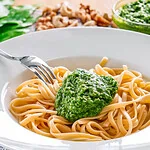 Ein Teller mit Spaghetti und einem Klecks grünem Bärlauch Pesto, garniert mit einer Gabel, die teilweise unter der Pasta verborgen ist, auf einem Tisch mit unscharfem Hintergrund.