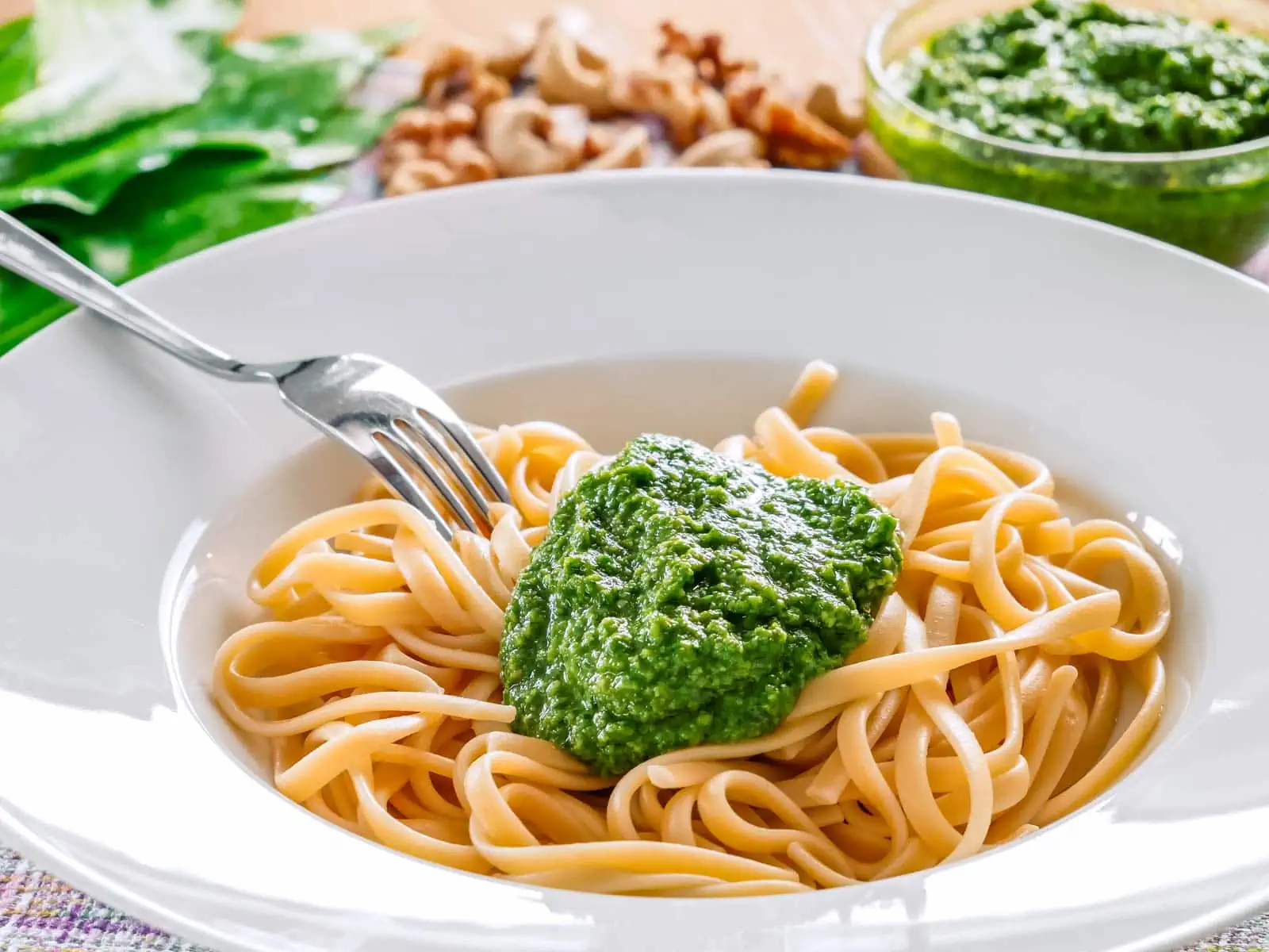 Ein Teller mit Spaghetti und einem Klecks grünem Bärlauch Pesto, garniert mit einer Gabel, die teilweise unter der Pasta verborgen ist, auf einem Tisch mit unscharfem Hintergrund.