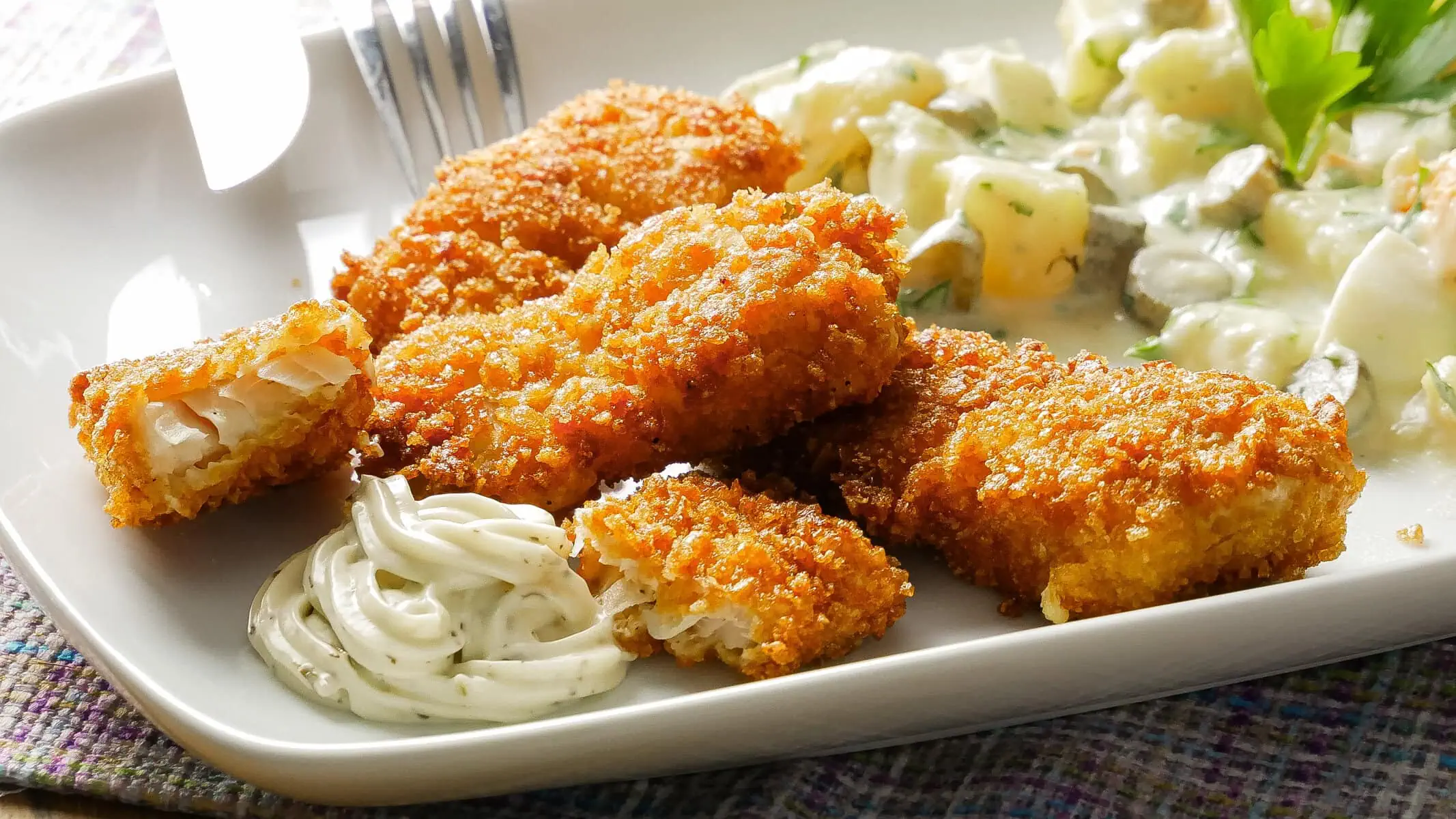 Fischstäbchen mit Cornflakes-Panade auf einem Teller, begleitet von Remoulade und Kartoffelsalat mit Gurkenwürfeln. Im Hintergrund eine Schale Cornflakes.