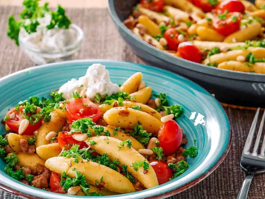 Eine Pfanne mit Schupfnudeln, Hackfleisch, Kirschtomaten und Kräutern, serviert auf einem blauen Teller mit einem Klecks Crème fraîche.