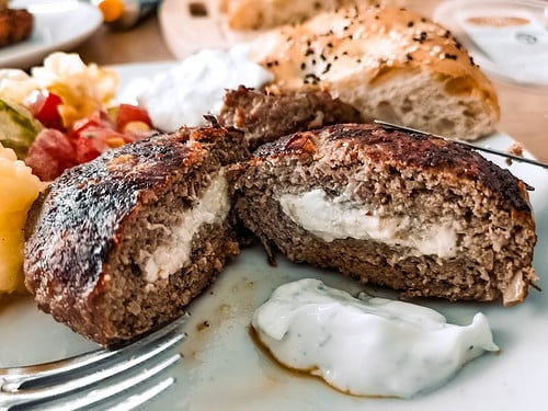 Aufgeschnittenes Bifteki, gefüllt mit Schafskäse, serviert mit Tsatsiki, frischem Salat und Fladenbrot im Hintergrund.