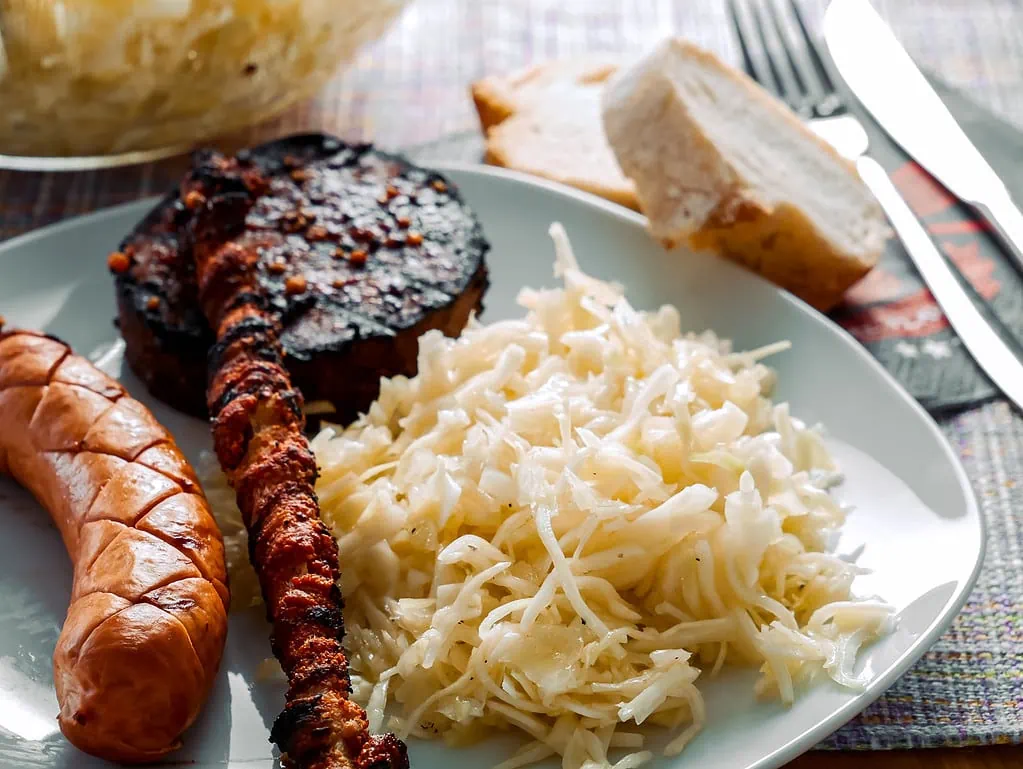 Ein Teller mit griechischem Krautsalat, gegrillter Wurst, Fleischspieß und gebratener Blutwurst, serviert mit frischem Brot im Hintergrund.