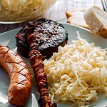 Ein Teller mit griechischem Krautsalat, gegrillter Wurst, Fleischspieß und gebratener Blutwurst, serviert mit frischem Brot im Hintergrund.