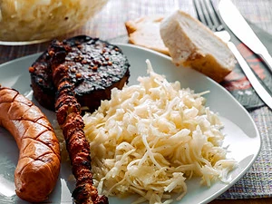 Ein Teller mit griechischem Krautsalat, gegrillter Wurst, Fleischspieß und gebratener Blutwurst, serviert mit frischem Brot im Hintergrund.