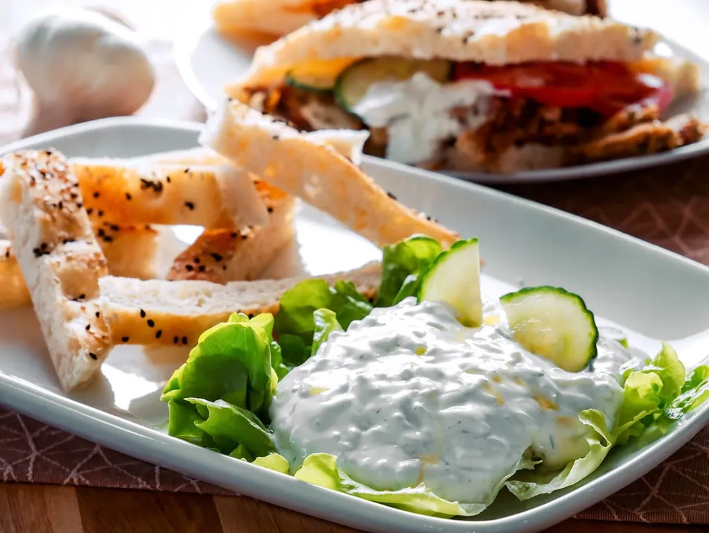 Ein Teller mit frischem Tsatsiki, garniert mit Gurkenscheiben und grünem Salat, serviert mit Sesam-Fladenbrotstreifen. Im Hintergrund ist ein gefülltes Fladenbrot mit Tomaten und Tsatsiki zu sehen.