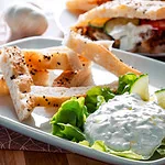 Ein Teller mit frischem Tsatsiki, garniert mit Gurkenscheiben und grünem Salat, serviert mit Sesam-Fladenbrotstreifen. Im Hintergrund ist ein gefülltes Fladenbrot mit Tomaten und Tsatsiki zu sehen.