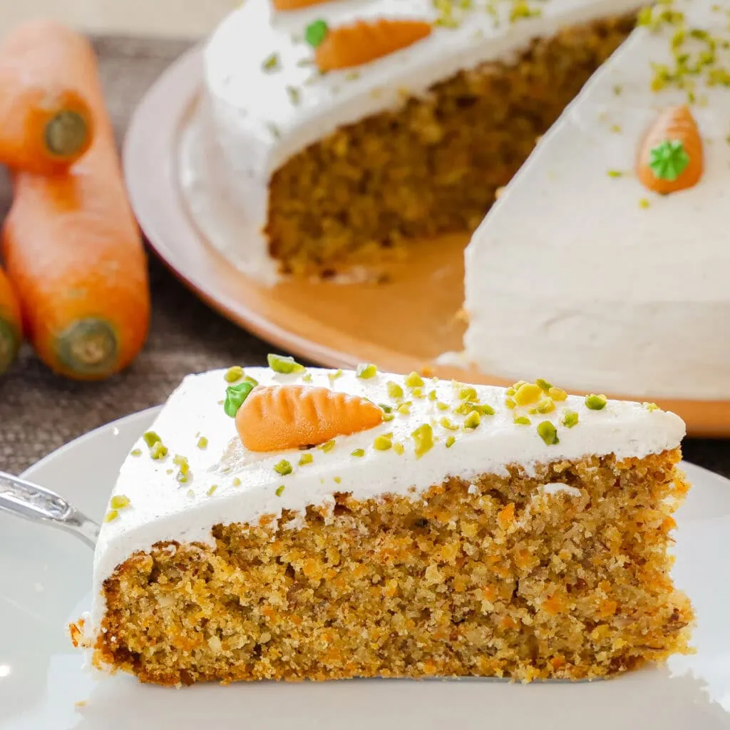 Ein Stück Karottenkuchen mit weißem Frischkäse-Frosting und Pistazien auf einem Teller, Karotten-Dekorationen im Hintergrund.