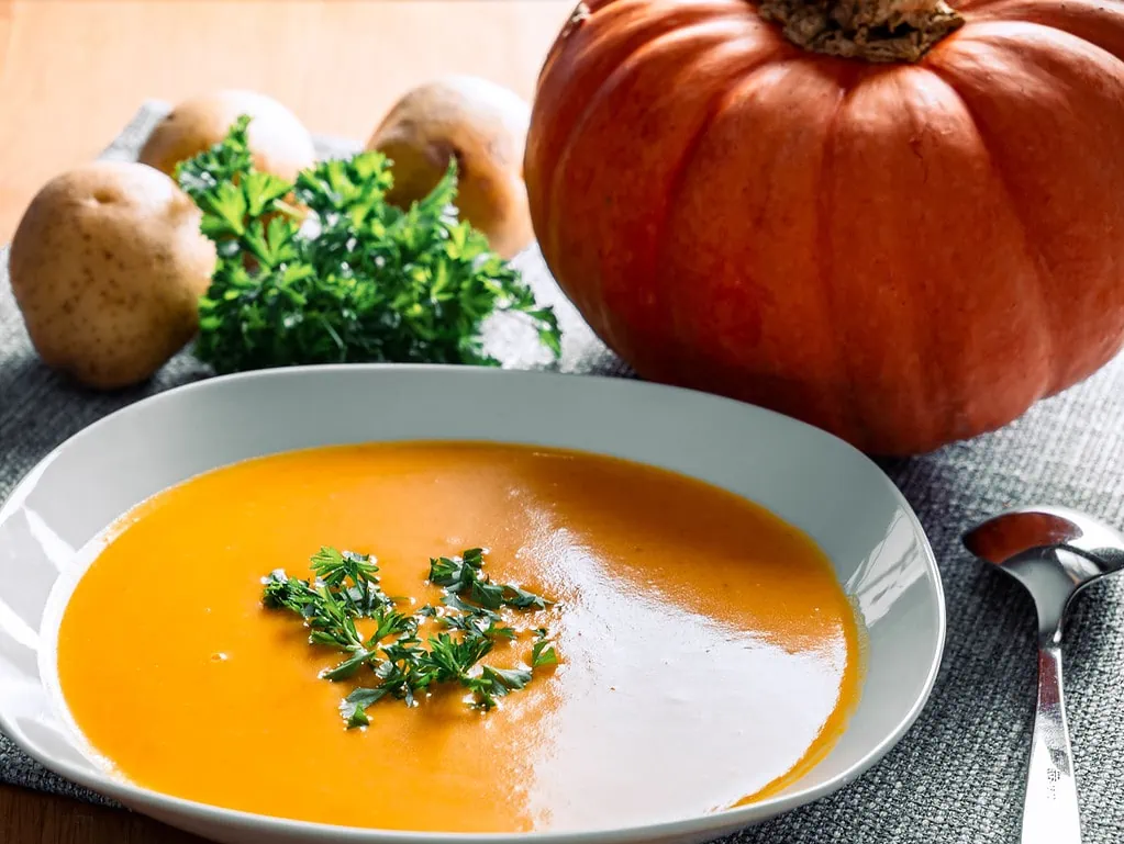 Ein Teller mit cremiger Kürbissuppe, garniert mit frischer Petersilie. Im Hintergrund liegen Kartoffeln, ein großer Kürbis und ein Bund Petersilie auf einer grauen Tischdecke. Ein Löffel liegt neben dem Teller.