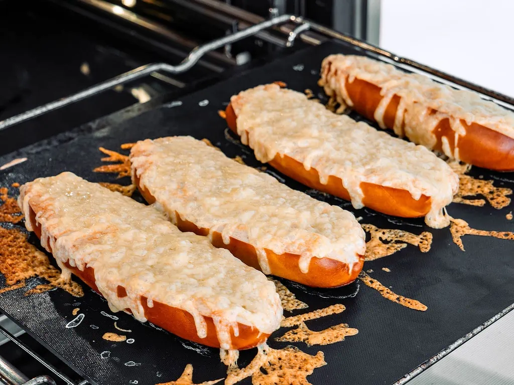 Drei frisch gebackene Käse-Laugenstangen auf einem Backblech im Ofen, überzogen mit geschmolzenem Käse.